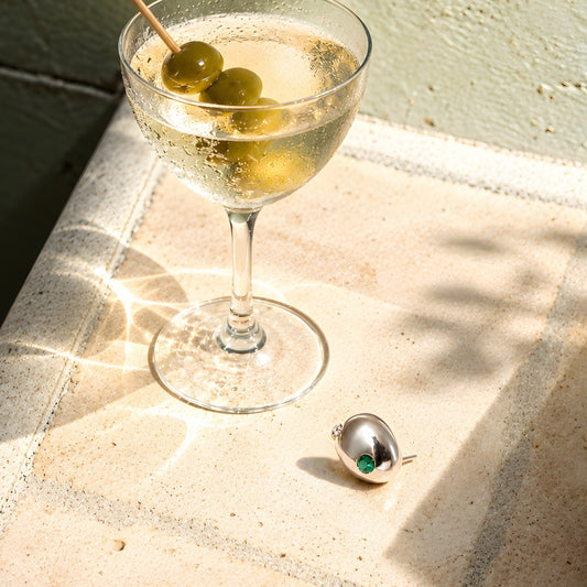 925 sterling silver pebble stud earring with green and clear zirconia beside a sunlit cocktail glass with olives.